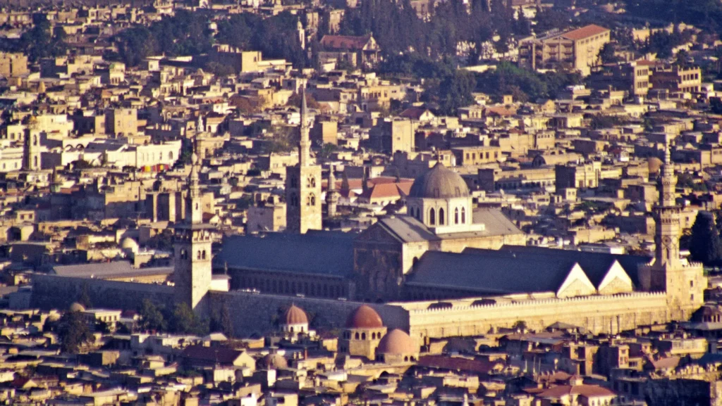 Die Große Umayyaden-Moschee in der Altstadt von Damaskus – Gesamtsicht von Nordwesten. Im Süden des umbauten Hofes die Gebetshalle mit Querhaus-Kuppel, an der linken bzw. rechten Ecke das „Jesus-Minarett“ und das „Qaytbay-Minarett“ sowie auf der Nordseite das „Minarett der Braut“